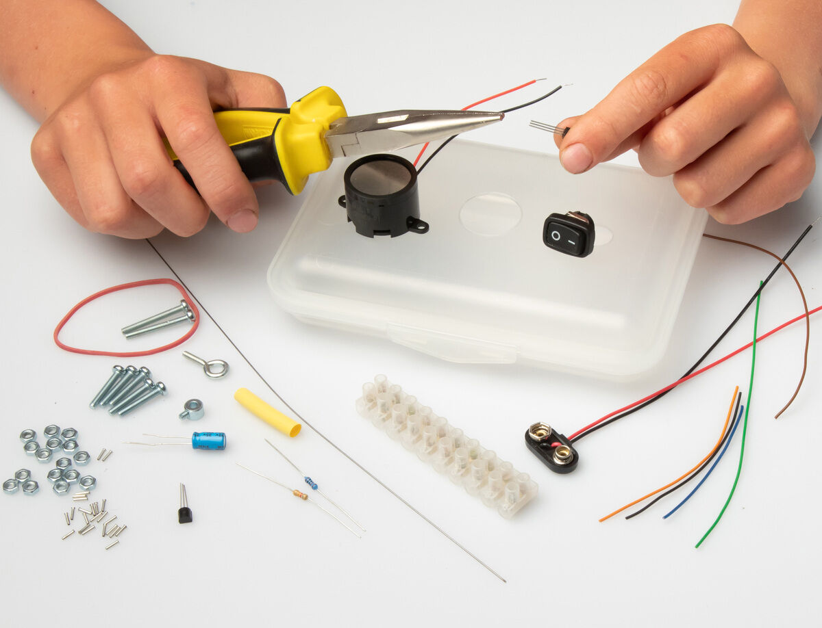 Ein Bild zeigt den Inhalt eines »Bewegungsalarm« Experimentierkastens. Eine Hand hält eine gelbe Zange und manipuliert eine Komponente. Verschiedene elektrische Bauteile wie Kabel, Schrauben, Muttern und ein Plastikgehäuse sind auf einem weißen Hintergrund verteilt.