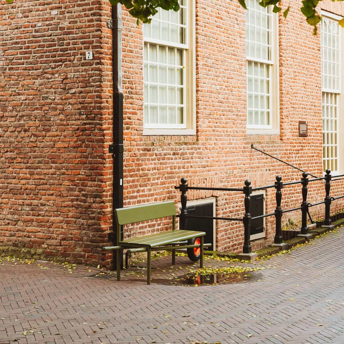 Vor einer roten Backsteinwand steht die grüne Schubkarrenbank »Wheelbench« von Weltevree. Das Möbelstück aus Holz und Metall ist von etwas Herbstlaub und nassem Kopfsteinpflaster umgeben. Links ranken sich grüne Blätter von einem Baum herab.
