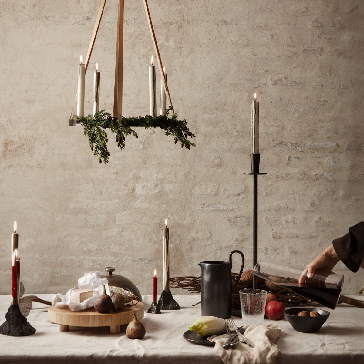 Ein stilvoller Adventskranz namens »Circle« hängt von der Decke. Der Kranz besteht aus Holz, dekoriert mit grünen Zweigen und weißen Kerzen. Auf einem Tisch darunter sind weitere Kerzen, Nüsse und ein Krug platziert. Eine Hand arrangiert die Szene. Die Farbpalette ist minimalistisch mit Holzfarben, Weiß, Rot, Grün und Schwarz.