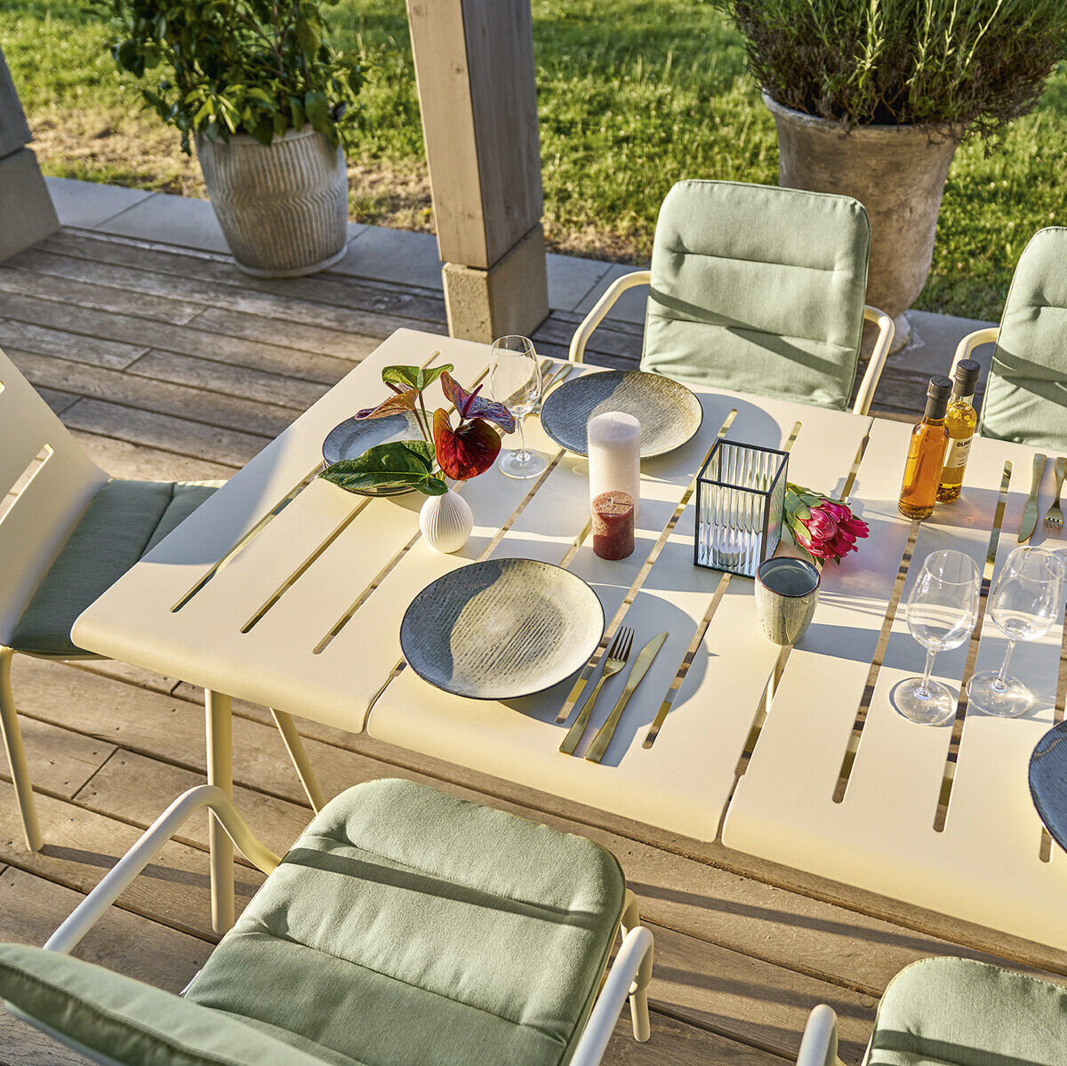 Ein beige-grauer Esstisch steht auf einer Holzterrasse, umgeben von je vier grünen und grauen Stühlen. Auf dem Tisch befinden sich verschiedene Gegenstände, darunter Teller, Gläser, Besteck, Blumen und Getränke. Im Hintergrund ist ein Garten mit grüner Wiese und Pflanzen zu sehen.