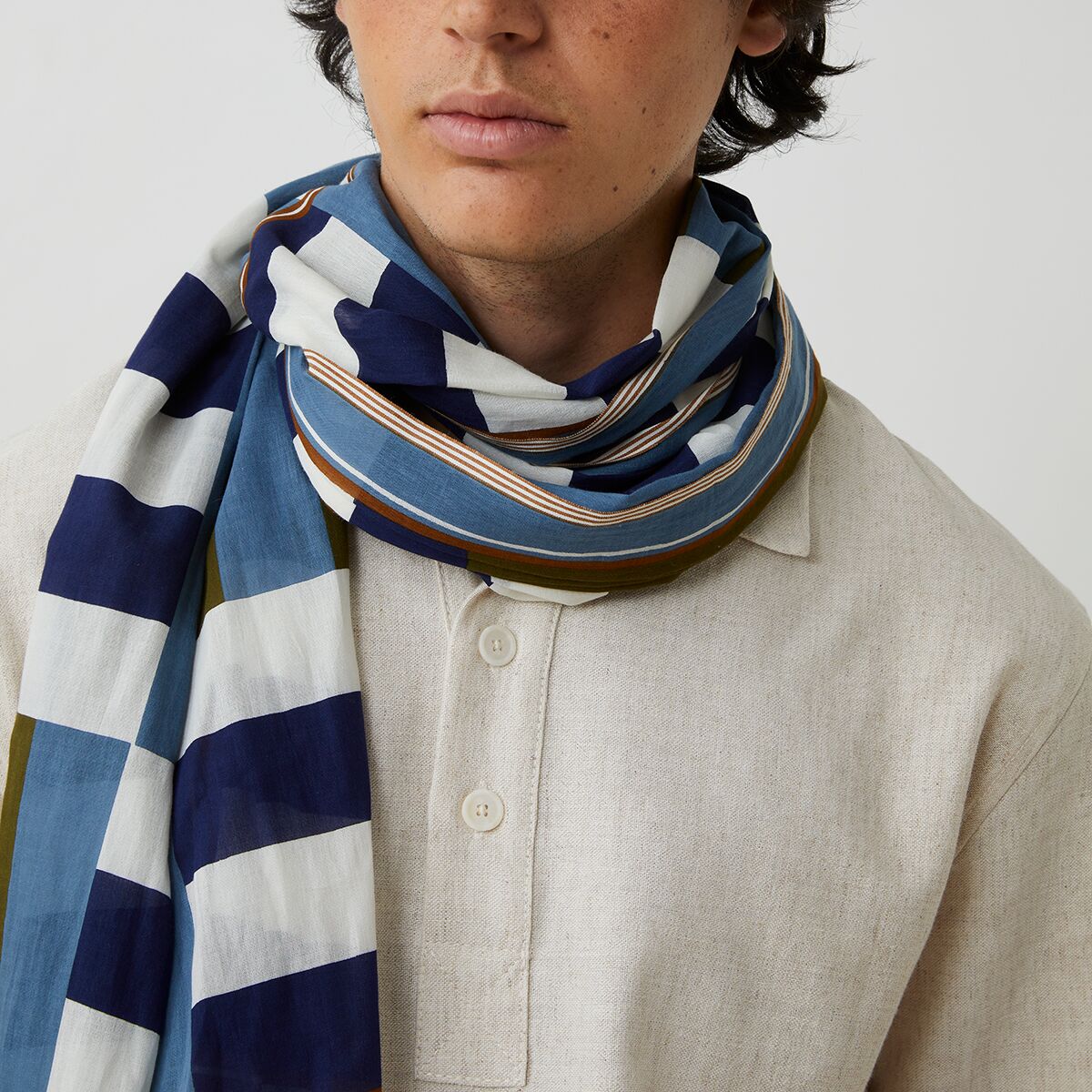 Ein Modell trägt einen großformatigen Baumwollschal No. 771 in Blau von MOISMONT, der breit blau-weiß gestreift und mit olivgrünen Akzenten versehen ist. Der Schal ist um den Hals gewickelt und hebt sich durch seine auffälligen Muster von einer beigen Leinenkleidung ab.