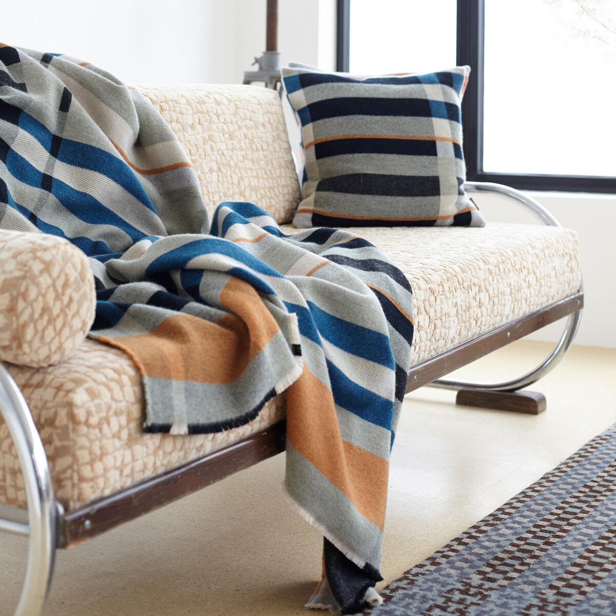 Ein elegantes Wohnzimmer mit einem beigen Sofa im skandinavischen Stil. Auf dem Sofa liegen eine gestreifte Decke mit Blau-, Grau-, Orange- und Beigetönen sowie ein passendes Kissen. Im Hintergrund befindet sich ein großes Fenster, das viel Licht in den Raum lässt.