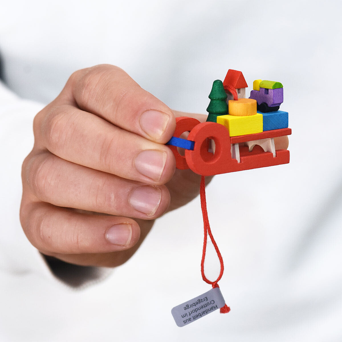 Eine Hand hält einen roten Holz-Weihnachtsbaumanhänger in Form eines Schlittens mit spielzeugartigen Figuren in verschiedenen Farben wie Grün, Gelb und Blau. Ein rotes Band und ein Etikett sind am Anhänger befestigt.