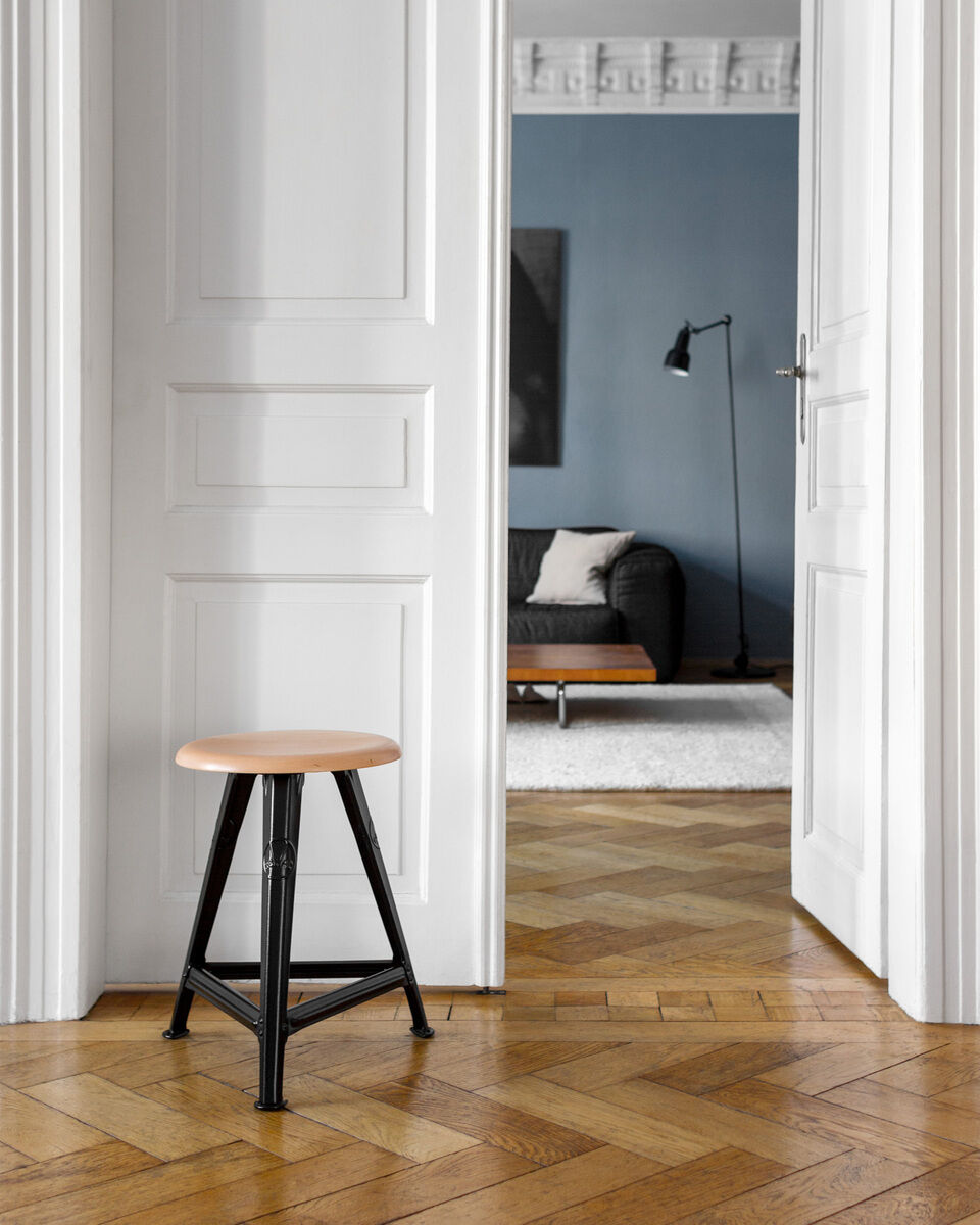 Ein stilvoller Schemel aus mattschwarzem Metall und hellem Eichenholz steht in einem eleganten Innenraum mit blau getünchten Wänden, einem schwarzen Sofa und einem weißen Teppich auf einem Fischgräten-Holzboden.