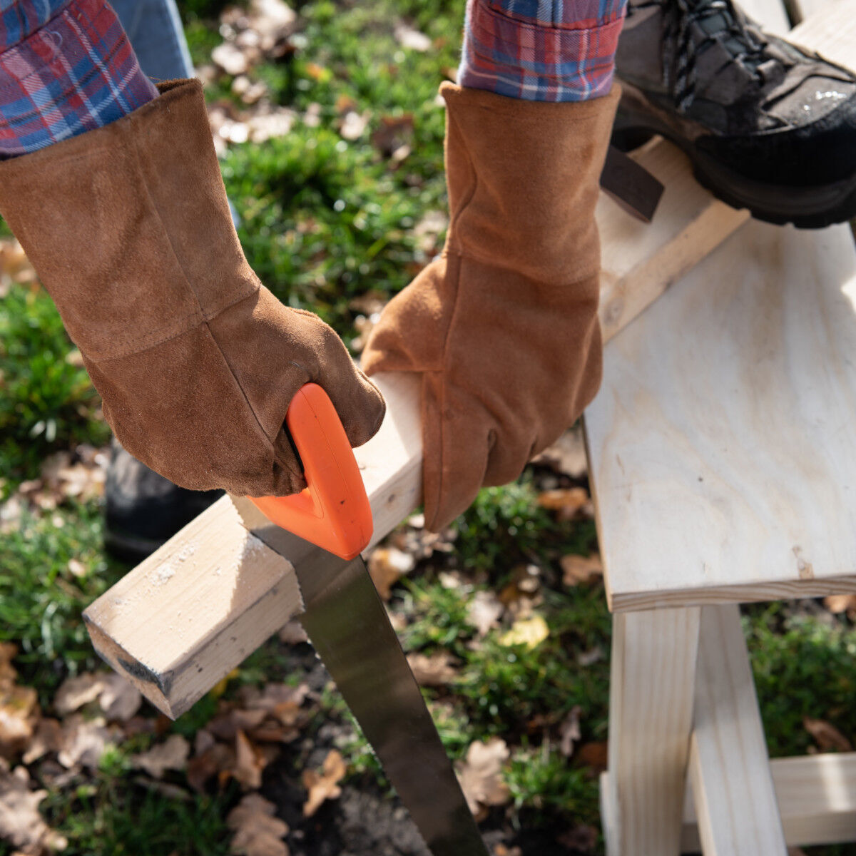 Person trägt braune Arbeitshandschuhe aus Rindleder und benutzt eine Handsäge, um ein Stück Holz draußen im Garten zu sägen.