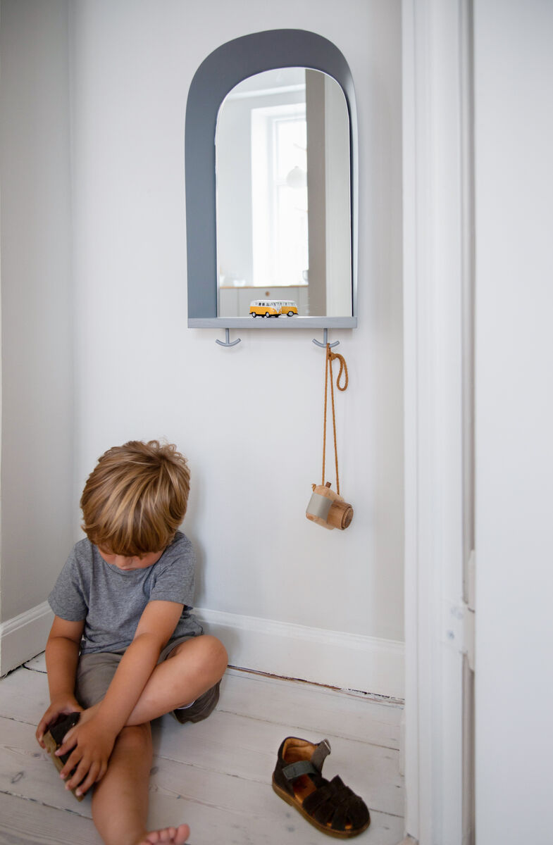 Ein Zimmer mit einem grauen, bogigen Spiegel an der Wand, unter dem zwei Haken und ein kleines Spielzeugauto platziert sind. Ein kleiner Junge sitzt auf dem Boden, trägt ein graues T-Shirt und blaue Shorts und spielt mit einem braunen Schuh. Der Fußboden ist aus hellem Holz.