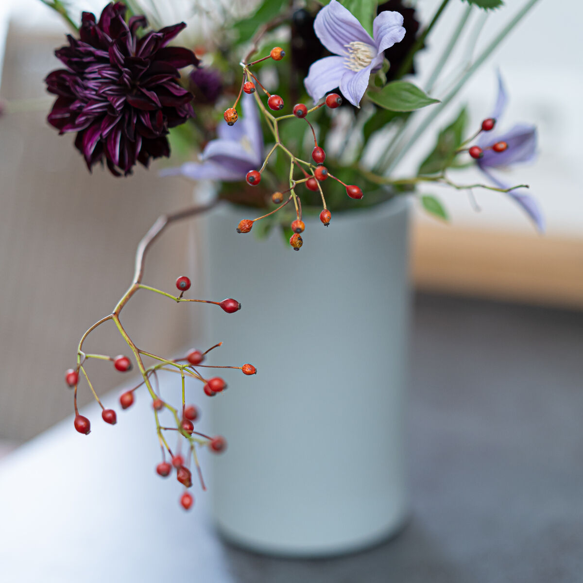 Porzellanvase in betongrün matt von Frau Caze, Modell "KULA" von Carolin Zeyher, steht auf einem Tisch. Gefüllt mit einem Blumenarrangement in Violett- und Schwarztönen sowie roten Beeren. Oberfläche matt, modernes schlichtes Design.