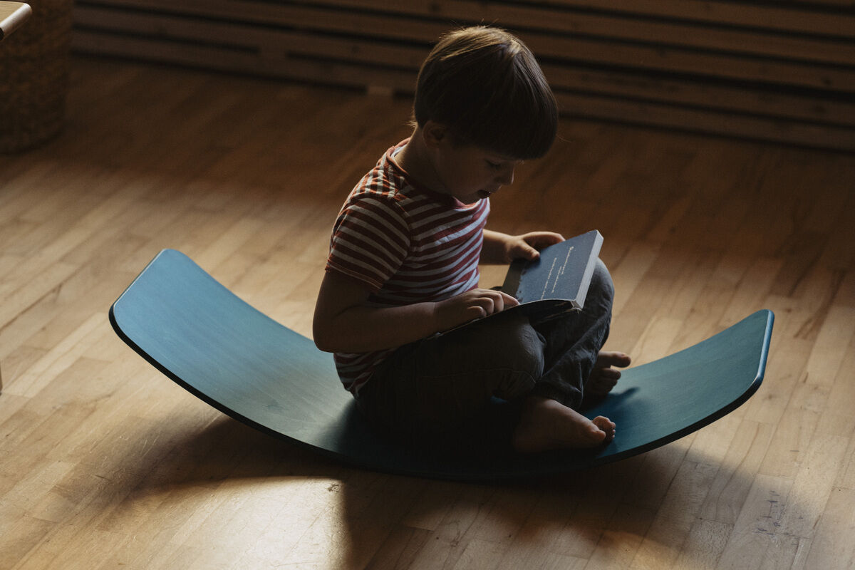 Ein Kind sitzt auf einem mintfarbenen Balance Board namens 'das.Brett' und liest ein Buch in einem abgedunkelten Raum mit Holzfußboden.