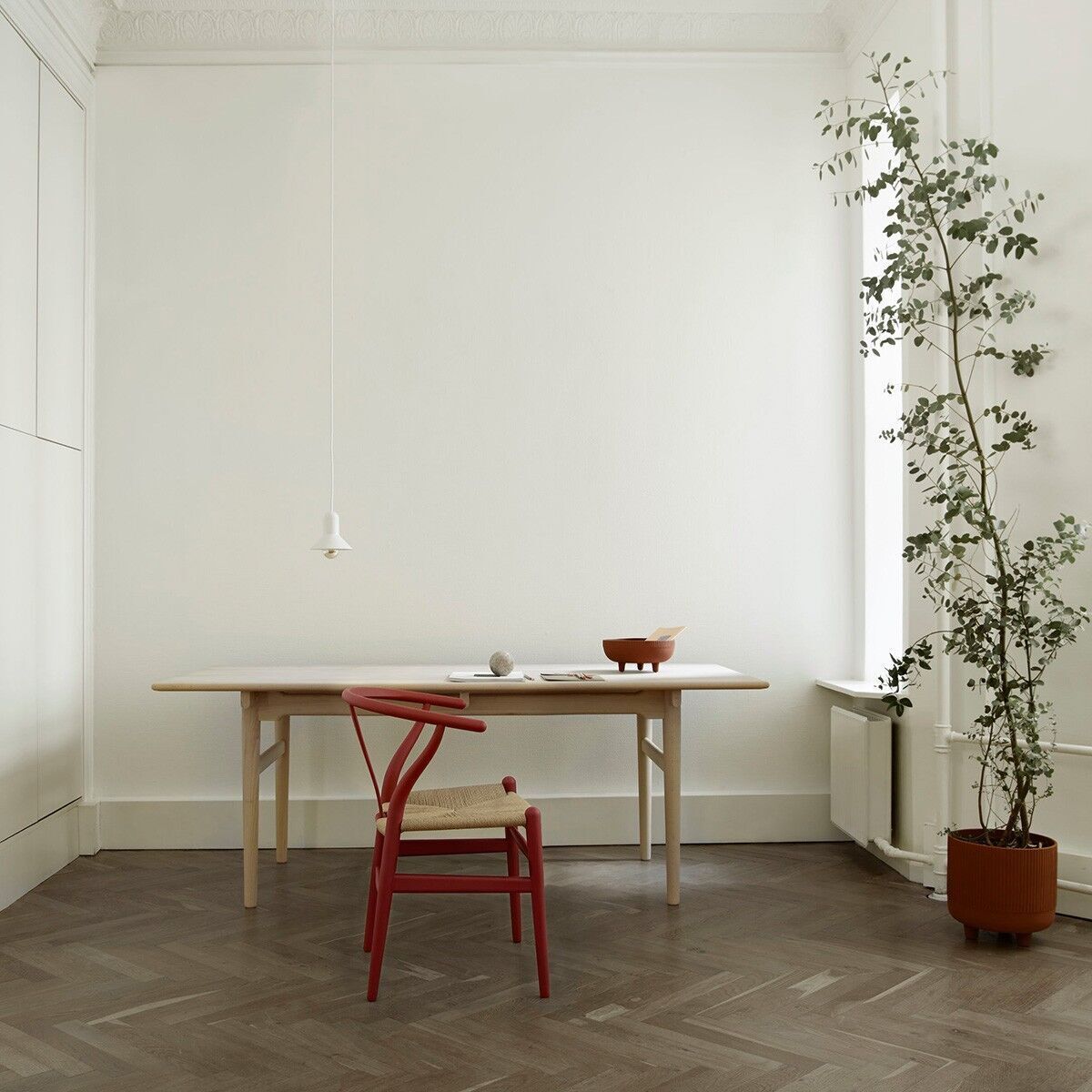 Ein minimalistisch eingerichtetes Zimmer mit einem eleganten Tisch und dem Designstuhl CH24 Soft von Carl Hansen in Rot. Eine grüne Pflanze steht in der Ecke und der Raum ist in hellen Tönen gehalten.