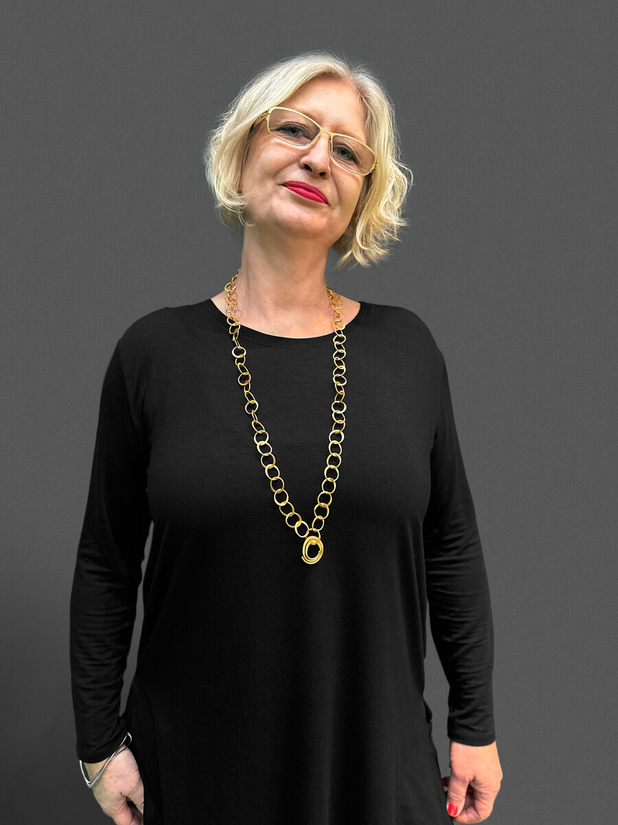 Eine Frau mit blonden Haaren und Brille trägt ein schwarzes einfaches Kleid und eine vergoldete silberne doppelte Halskette mittlerer Länge. Der Hintergrund ist grau.