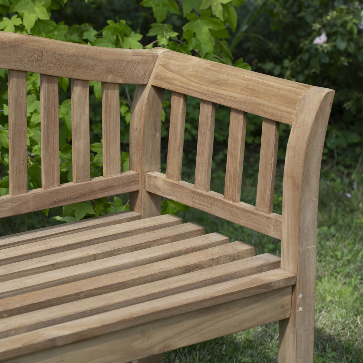 Detailaufnahme einer stilvollen Teakholz-Gartenbank »Wave« von bene living. Die Bank steht auf grünem Rasen vor üppigen grünen Sträuchern. Das geschwungene Rückenteil und die Armlehne sind aus naturfarbenem Holz mit markanter Maserung gefertigt.