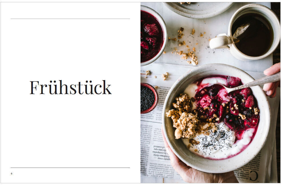 Ein aufgeschlagenes Kochbuch mit dem Titel 'Frühstück' befindet sich rechts im Bild und zeigt ein Rezept für ein gesundes Frühstück bestehend aus Joghurt, Müsli und frischen roten Beeren, begleitet von einer Tasse Tee. Die Seite auf der linken Seite des Buches ist nur textlich gestaltet.