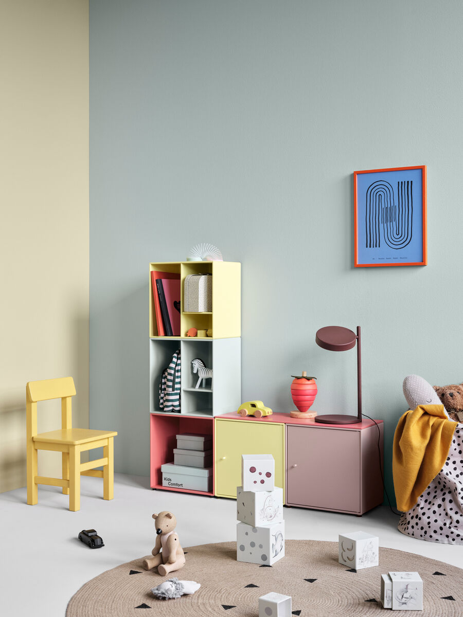 Ein farbenfrohes Kinderzimmer mit einem Regal namens 'Montana Mini 1002', gefüllt mit Spielzeug und Büchern, neben einem kleinen gelben Stuhl. Unter dem Regal befindet sich eine rote Lampe. An der Wand hängt ein blaues Bild. Der Raum ist in sanften Pastelltönen gestrichen.