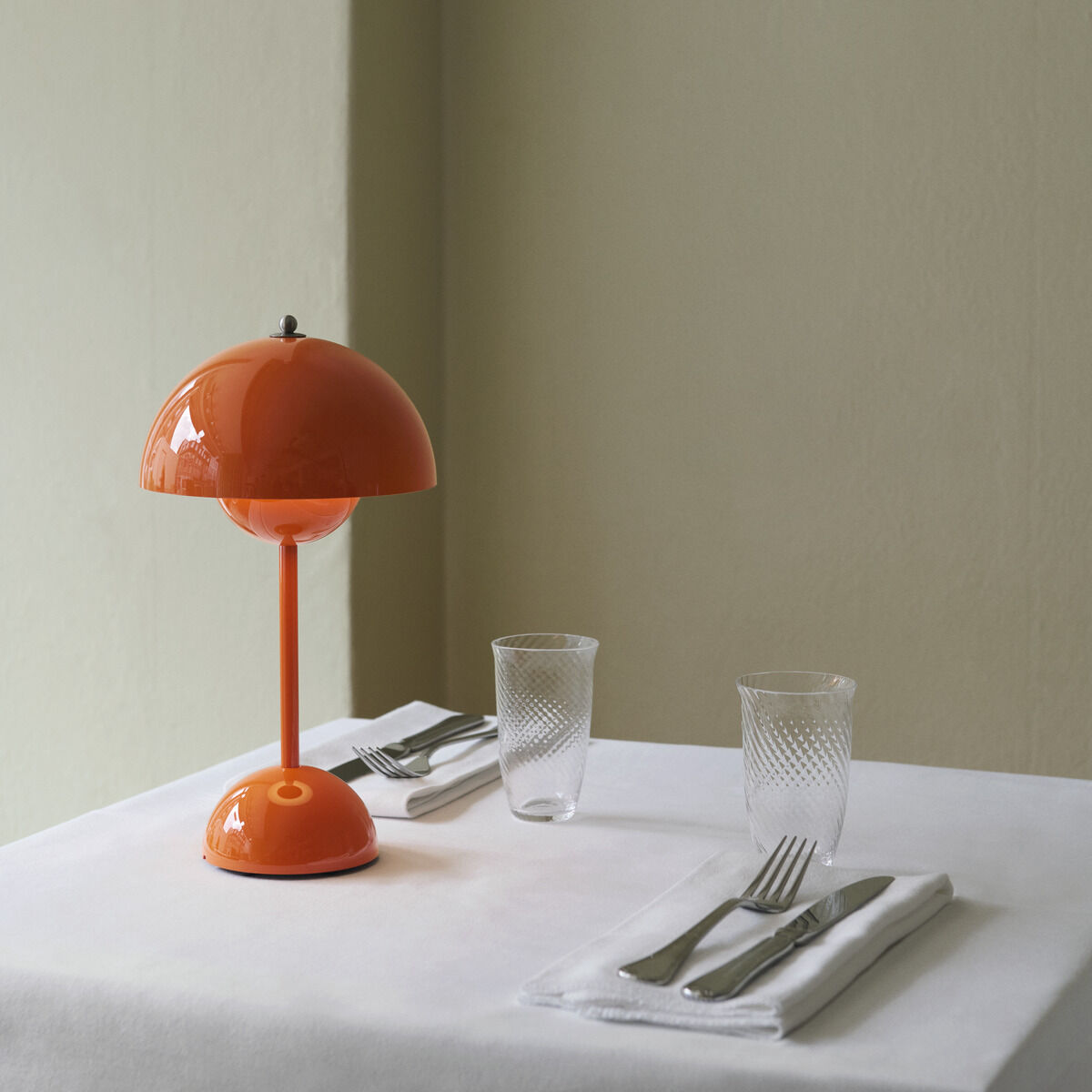 Auf einem weiß gedeckten Tisch stehen zwei Gedecke mit Messer, Gabel und Glas sowie eine leuchtend orangefarbene, dimmbare Tischleuchte Flowerpot VP9 von &Tradition in Retro-Design. Die Wand im Hintergrund ist hellbeige, ein Holzstuhl teilweise sichtbar.