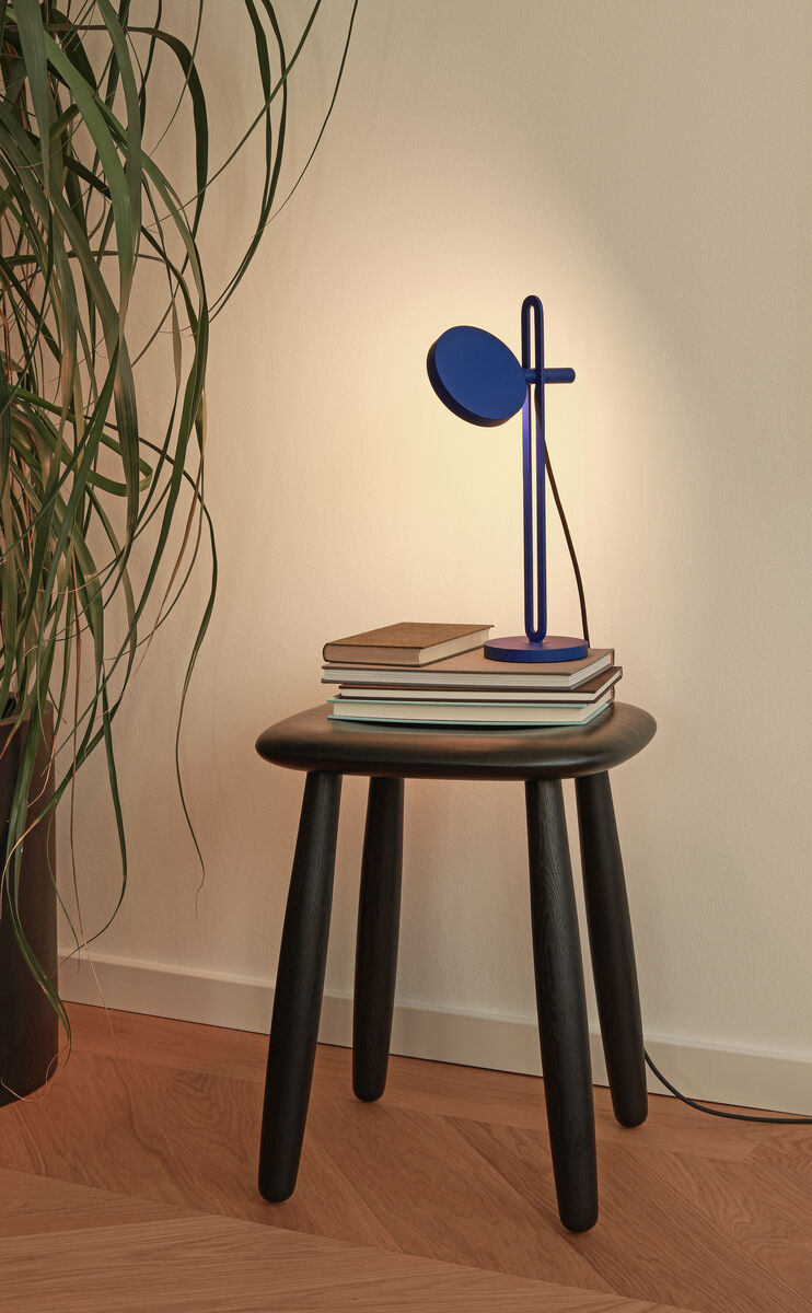 Eine Tischlampe ‚Echo’ steht auf einem kleinen, schwarzen Holztisch. Daneben liegen Bücher. Im Hintergrund ist eine grüne Pflanze vor einer cremefarbenen Wand und einen Holzboden zu sehen.