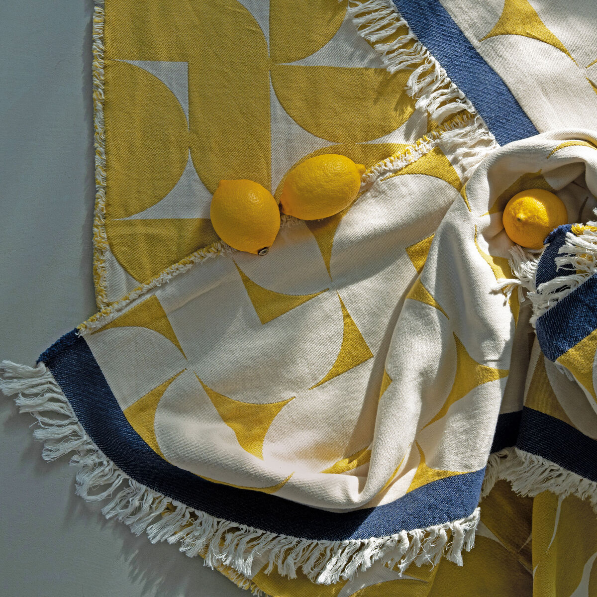 Gelb-weiß-gemusterte Baumwolldecke mit Fransen, drapiert mit drei Zitronen darauf, in hellem und modernem skandinavischem Design.