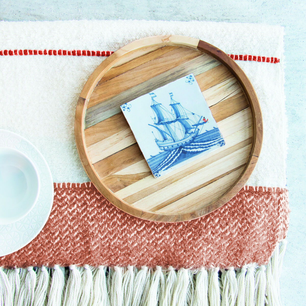 Eine Keramikfliese mit einem blauen Schiffsmotiv liegt auf einem hölzernen Serviertablett. Daneben befindet sich eine weiße Schüssel auf einer rot-weißen Textildecke.