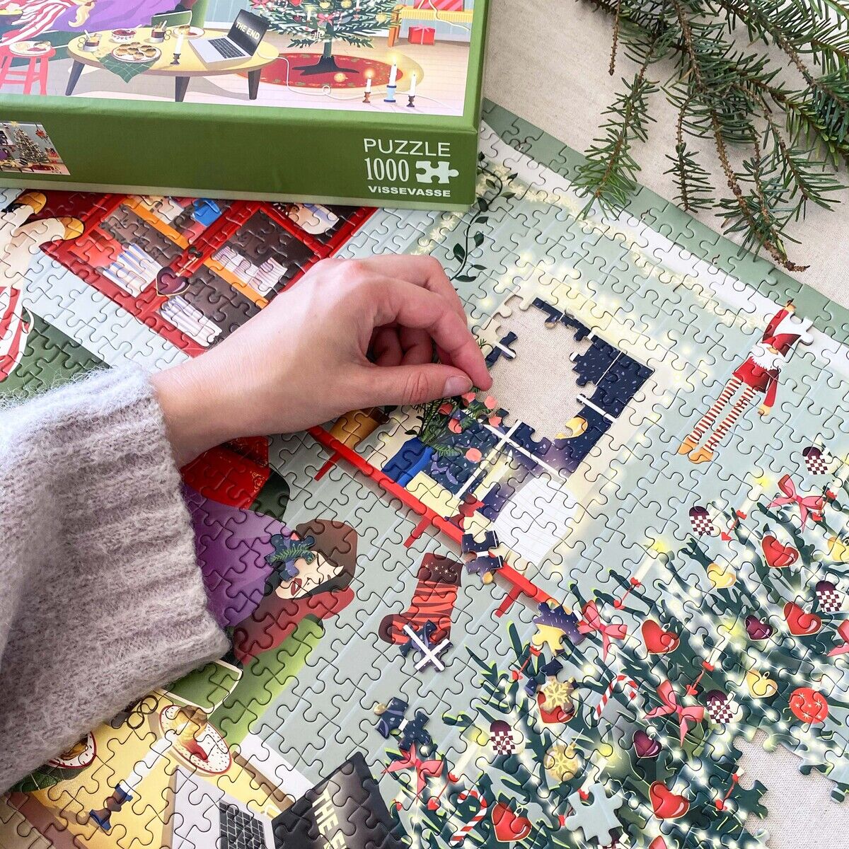 Eine Person legt das 1000-Teile-Puzzle »Faule Tage« von ViSSEVASSE. Das Motiv zeigt ein festlich geschmücktes Wohnzimmer mit Weihnachtsbaum, Geschenken und Figuren. Auf dem Tisch liegen Tannenzweige. Im Hintergrund ist der Karton des Puzzles zu sehen.