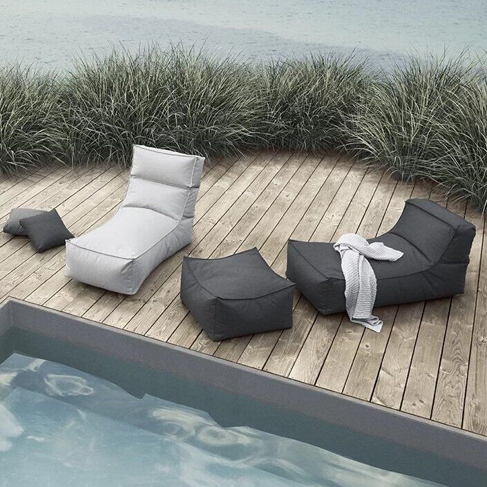 Moderner Outdoor Lounger 'STAY' auf einer Holzterrasse neben einem Pool, umgeben von Grünen Sträuchern. Die Möbel bestehen aus grauen und weißen Lounge-Sesseln und einem Beistelltisch.