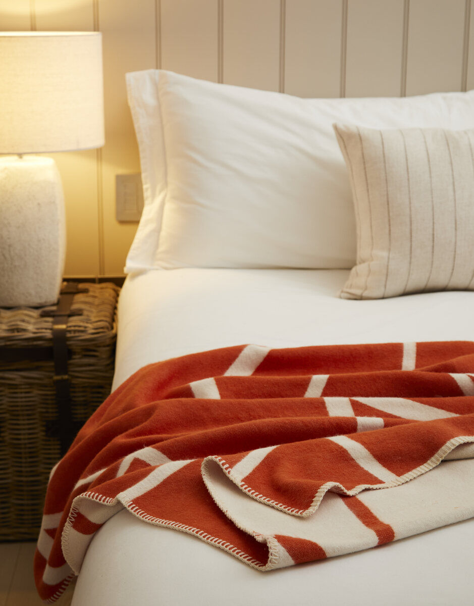 Ein gemütlich eingerichtetes Schlafzimmer mit einer Zimtrot-weiß gestreiften Baumwolldecke von Twig, die kunstvoll auf einem weißen Bett arrangiert ist. Ein Kissen mit Streifenmuster in Naturfarben und eine geflochtene Aufbewahrungskiste sind ebenfalls im Bild zu sehen.