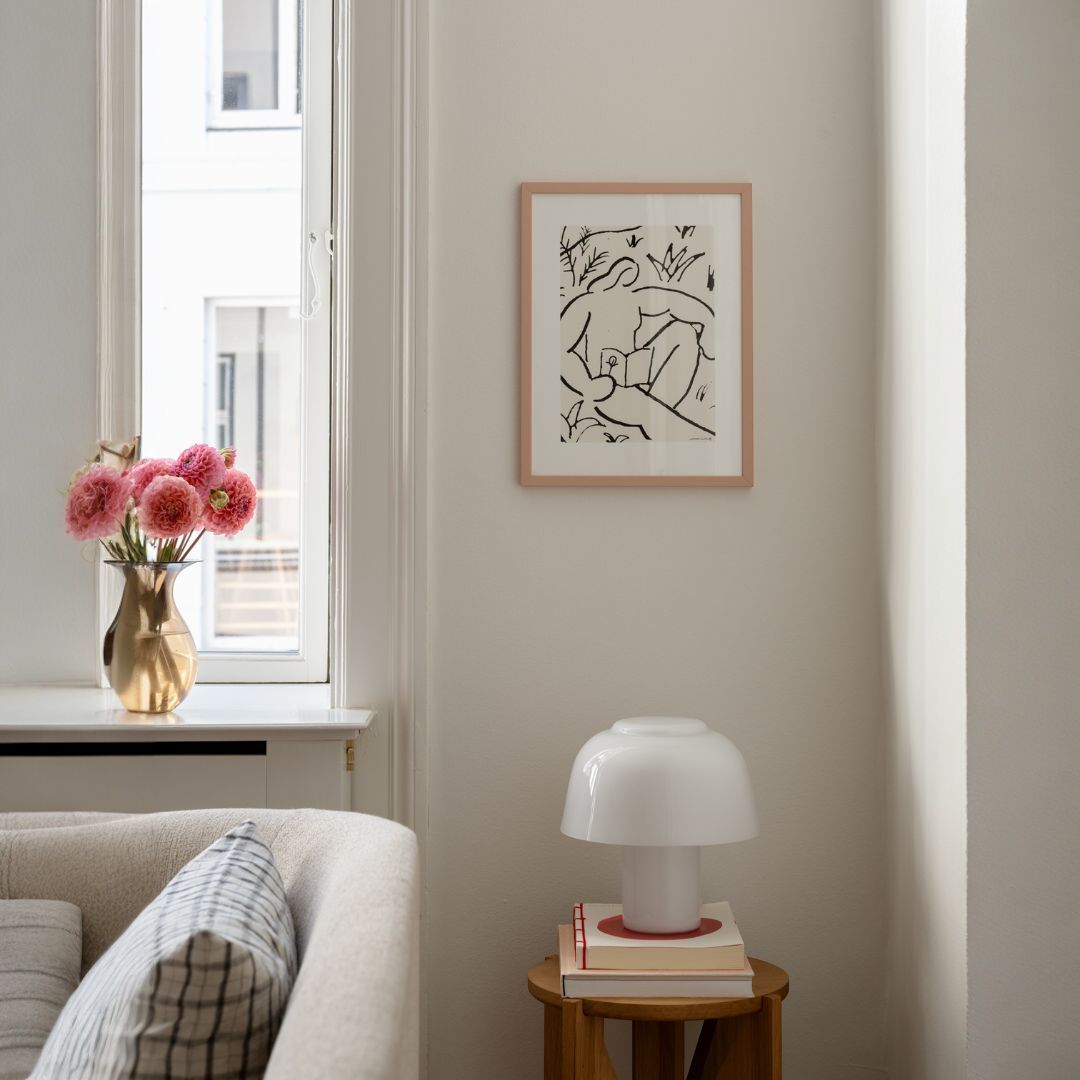 Ein minimalistisches Kunstwerk mit dem Titel 'The Reader', gerahmt und an einer weißen Wand aufgehängt. Daneben steht auf einem kleinen Tisch eine weiße Lampe, gestapelte Bücher, und ein Sofa mit Kissen. Ein Fenster mit Blumen in einer Vase sorgt für Tageslicht.