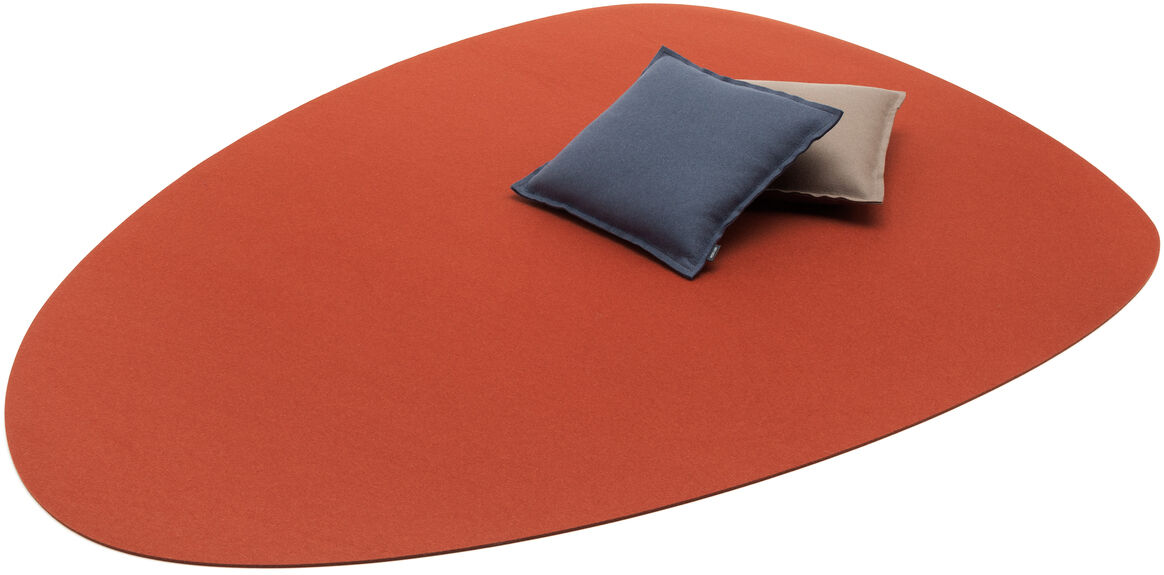 Ein roter Teppich »Stone« mit unregelmäßiger ovaler Form. Zwei Kissen, eines in Blau und eines in Beige, liegen darauf.