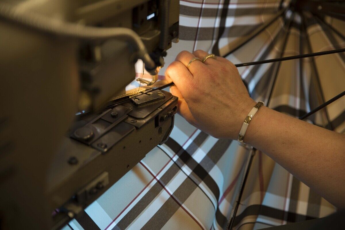 Eine Nahaufnahme einer Hand, die an einer Nähmaschine arbeitet, während sie an einem Schirm mit einem Ledergriff und einem karierten Muster näht.