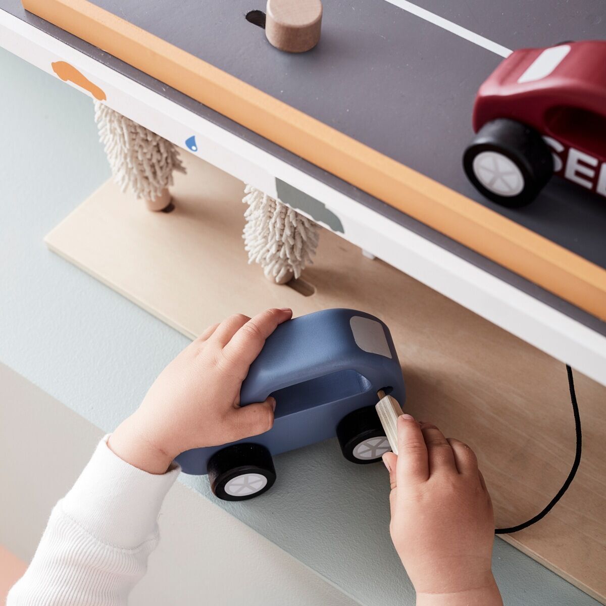 Ein Kind spielt mit einer aus Holz gefertigten Autowerkstatt. Hände eines Kindes halten ein blaues Holzauto, während ein rotes Auto auf einer grauen Spielfläche steht. Die Werkstatt besteht aus natürlichem Holz und enthält spielerische Details.