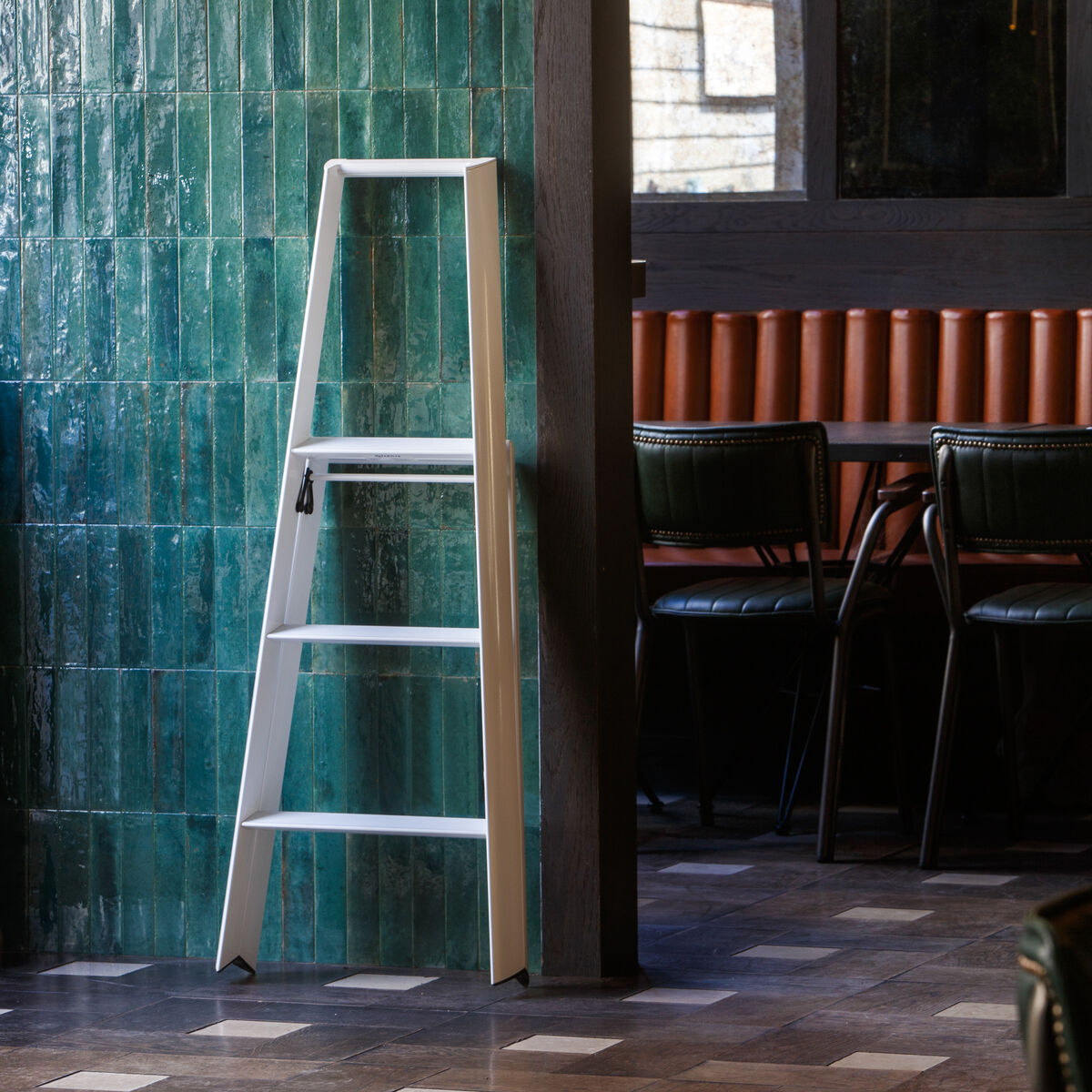 Eine moderne, dreistufige Klapptrittleiter aus weißem Aluminium steht an einer Wand mit glasierten, grünen Fliesen in einem stilvoll eingerichteten Innenraum. Im Hintergrund sind schwarze Stühle und braune Sitzpolster zu sehen. Die Umgebung wirkt wohnlich und modern.