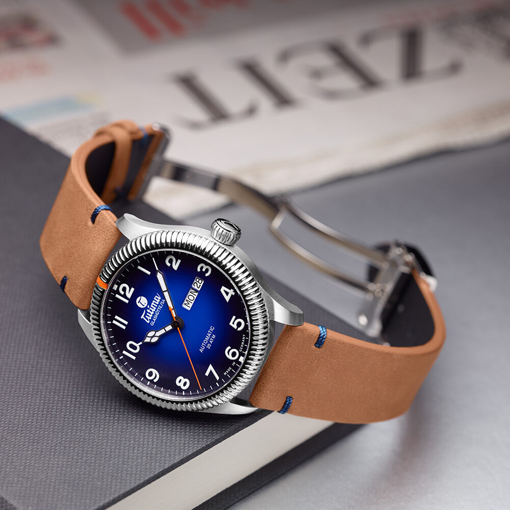 Die Tutima Glashütte Grand Flieger Classic Automatic ZEIT-Sonderedition ist auf einer Zeitung mit großem 'ZEIT'-Schriftzug drapiert. Sie zeigt ein glänzend blaues Zifferblatt mit orangefarbener Sekunde, Edelstahlgehäuse, cognacfarbenem Lederarmband mit blauer Ziernaht und Faltschließe.