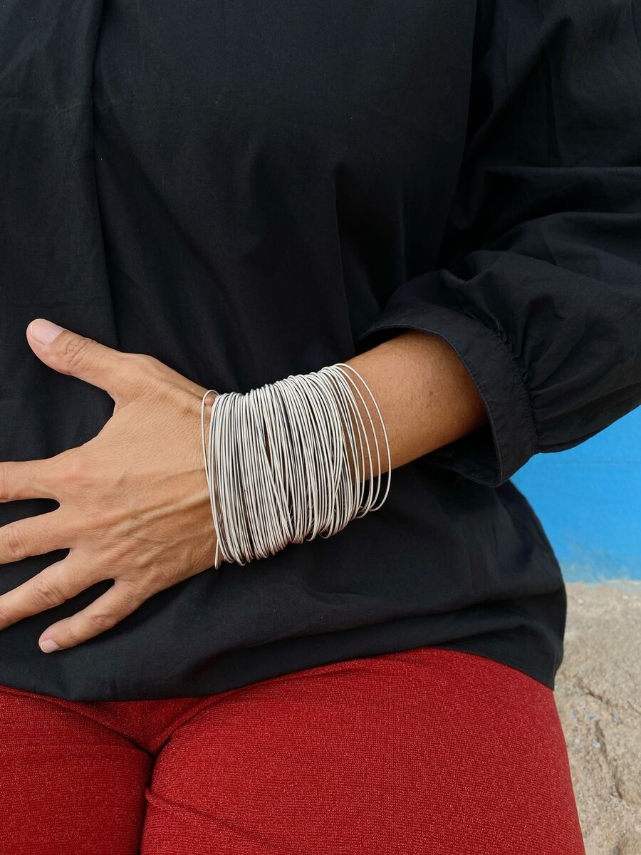 Eine Hand trägt das Armband »97« aus Edelstahlfedern, kombiniert mit einer schwarzen Bluse und roten Hosen vor einem blauen Hintergrund.