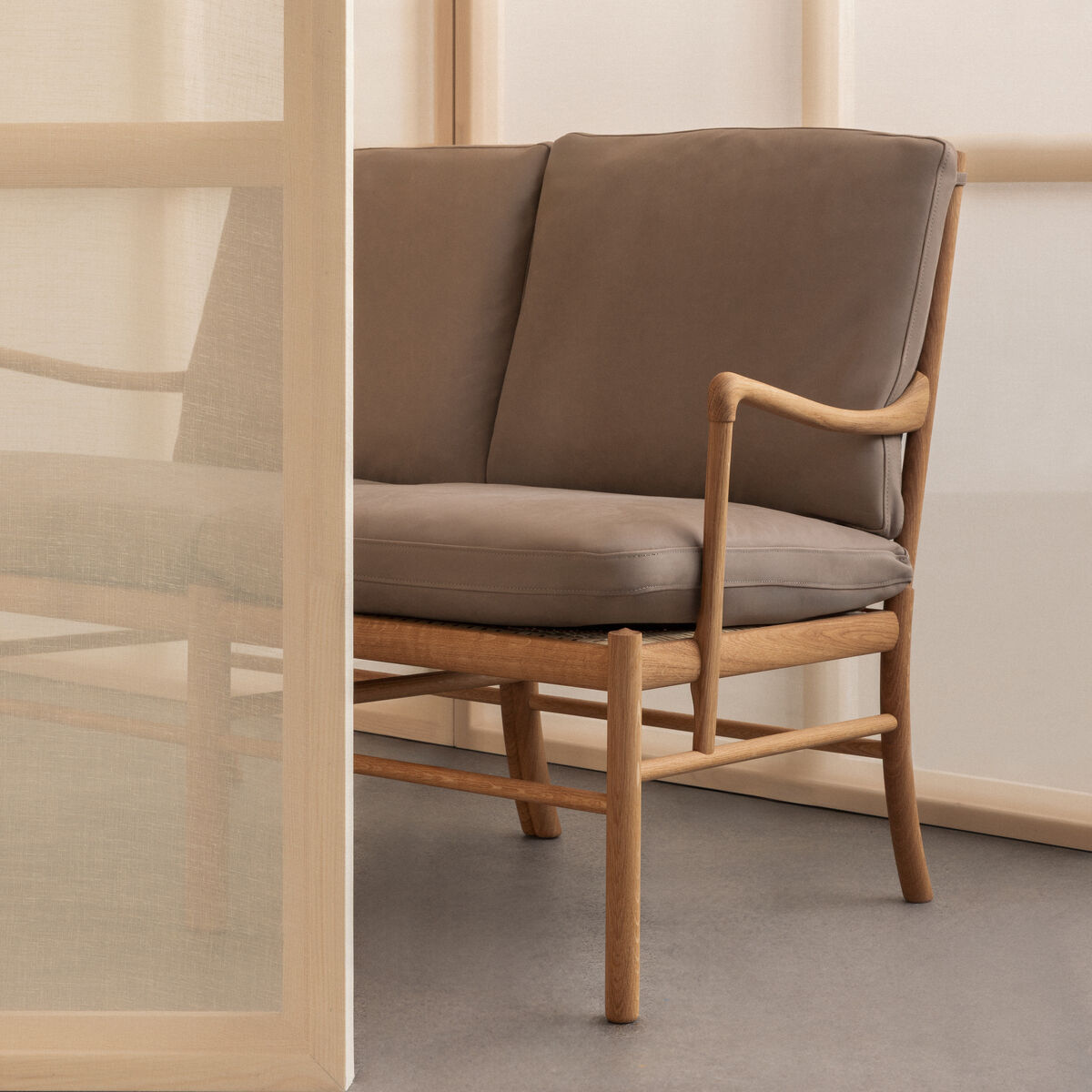 Eleganter Sessel mit hellbraunem Holzrahmen und braunen Kissen steht in einer minimalistischen, modernen Umgebung mit neutralen Wand- und Bodentönen. Der Sessel hat sanft geschwungene Armlehnen und klare Linien.