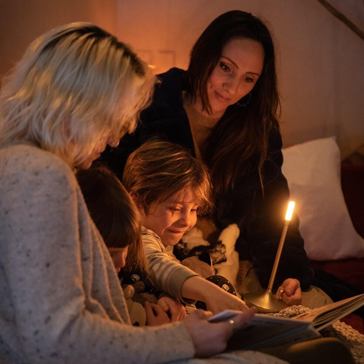 Eine Familie mit zwei Kindern sitzt in einem gemütlichen Wohnzimmer. Eine Wick Tischlampe leuchtet warm und erhellt die Szene. Die Mutter liest den Kindern vor, die aufmerksam zuhören.