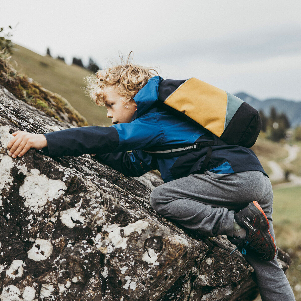 Ein Kind klettert auf einen Felsblock in einer bergigen Landschaft. Es trägt eine zweifarbige Tasche »Popoyo« in Blau und Gelb.