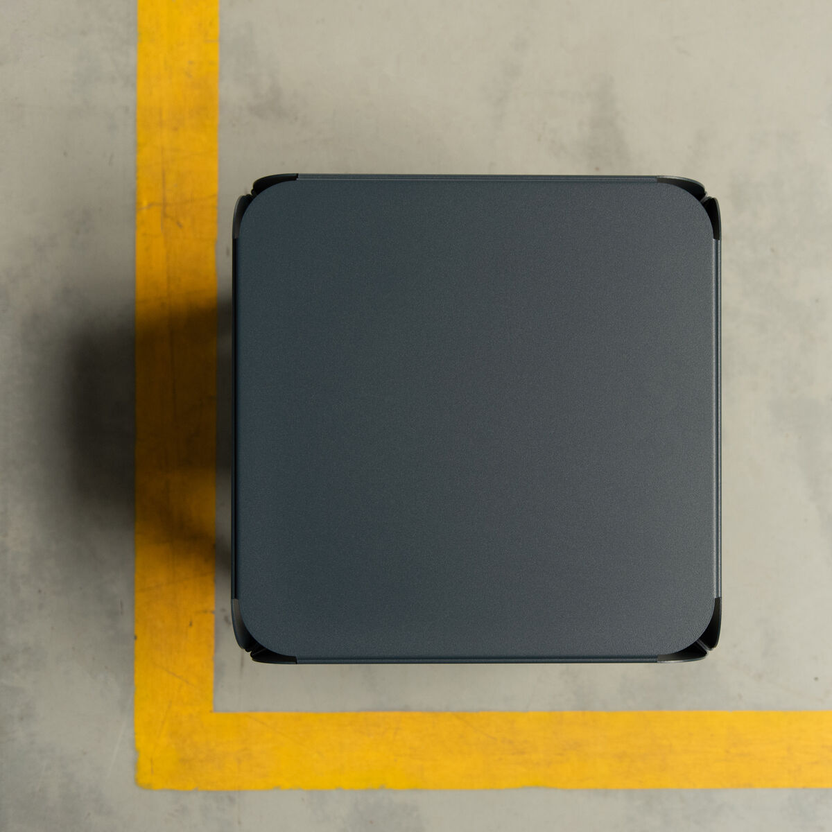 Ein quadratischer Hocker mit minimalistischer, moderner Gestaltung in einem dunklen Farbton. Er steht auf einem Boden mit hellgrauer Oberfläche und einem markanten gelben Streifen.