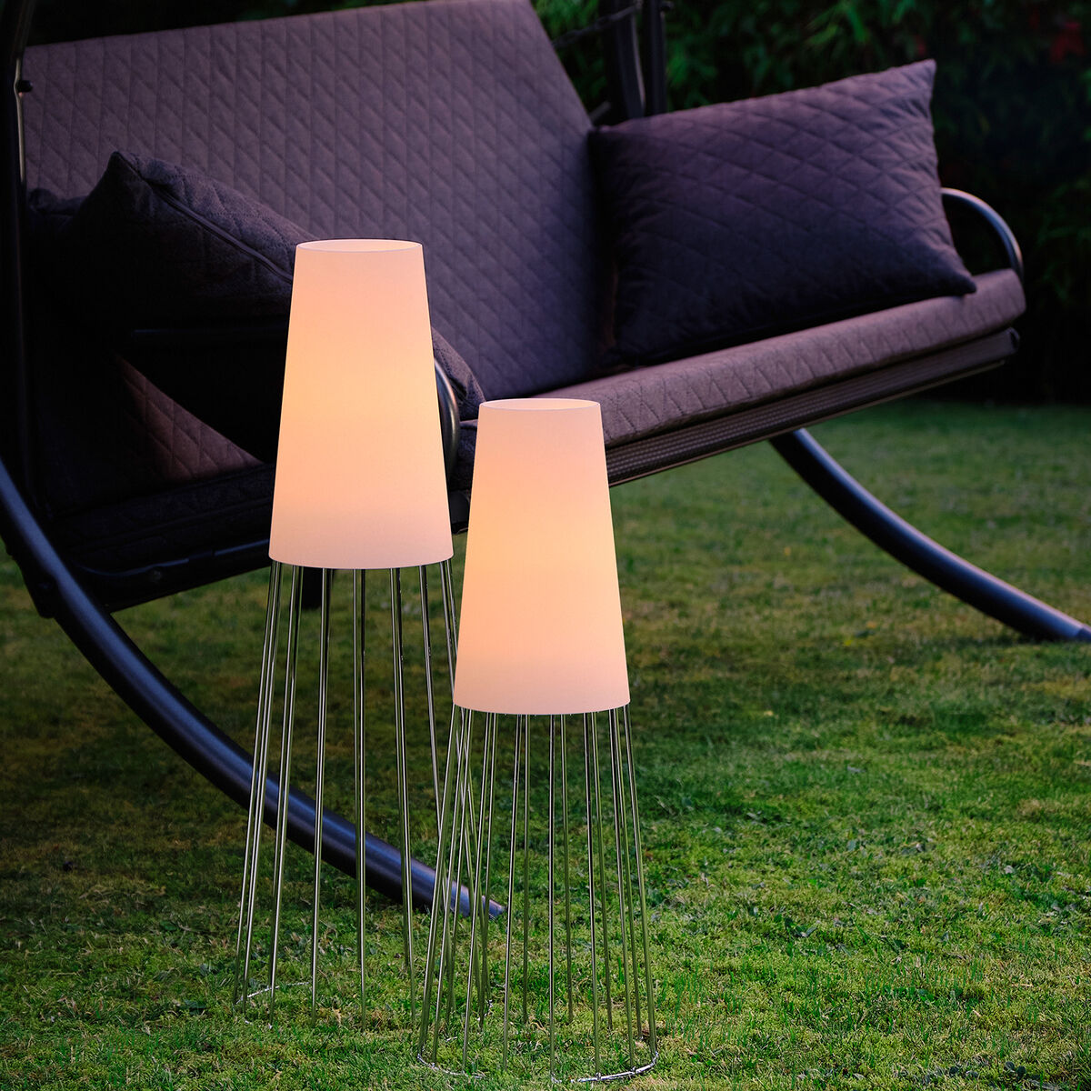 Zwei Gartenlichter vom Typ 'Lighthouse' leuchten neben einer dunklen Gartenschaukel auf einem grünem Rasen, umgeben von Bäumen und Sträuchern.