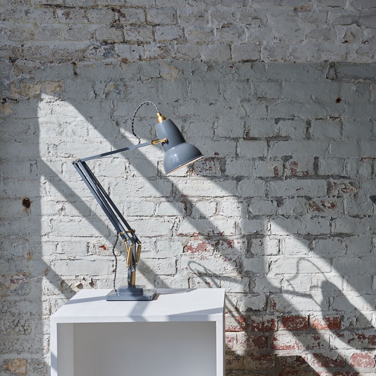 Die Tischleuchte »Anglepoise Original 1227 Brass Desk Lamp« in Grau und Messing steht auf einem weißen Tisch vor einer gemauerten Wand. Die Schreibtischlampe hat ein industrielles Design und ist verstellbar.