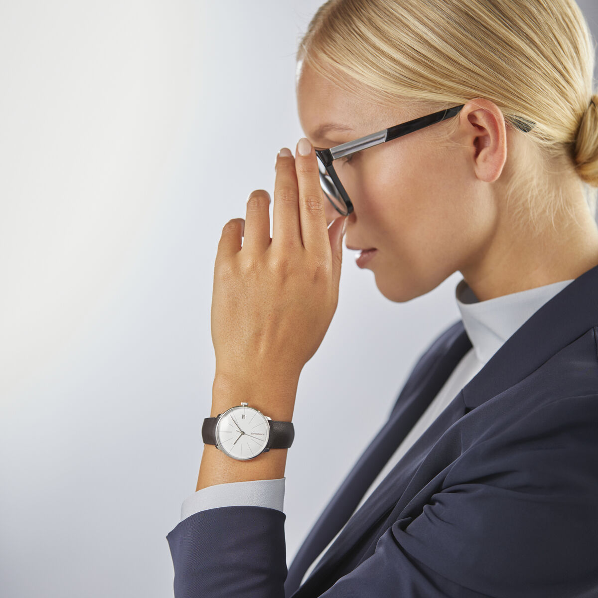 Eine elegante Frau mit blonden Haaren trägt eine schwarze Brille und eine blau-weiße Anzugjacke. Sie trägt die ZEIT-Sonderedition »Meister fein Kleine Automatic« von Junghans am Handgelenk, eine elegante, minimalistisches Uhr in Schwarz und Weiß.