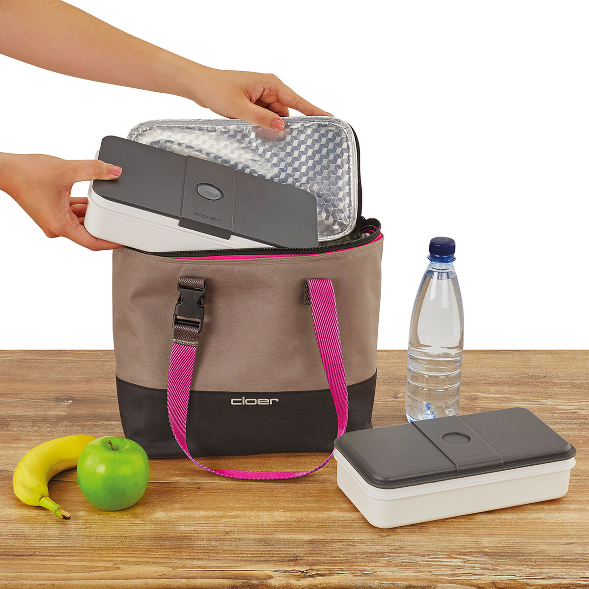 Lunch-Tasche mit rosa Griff auf einem Tisch, gefüllt mit einer Lunchbox. Eine Wasserflasche, eine weitere Lunchbox, ein grüner Apfel und eine Banane liegen daneben. Eine Hand holt eine Lunchbox aus der Tasche.