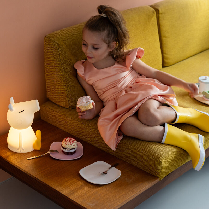 Ein kleines Mädchen in einem rosa Kleid und gelben Stiefeln sitzt auf einem gelben Sofa. Sie hält einen Muffin in der Hand und neben ihr auf einem Holztisch befindet sich ein leuchtendes Nachtlicht in Form der Comic-Figur Peppa Pig.