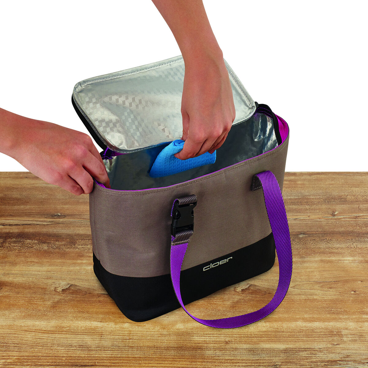 Eine Lunch-Tasche mit beige-schwarter Außenhülle und violetten Tragegriffen. Die Tasche wird von zwei Händen geöffnet und enthält ein im Inneren liegendes Kühlpack. Sie befindet sich auf einem Holztisch.