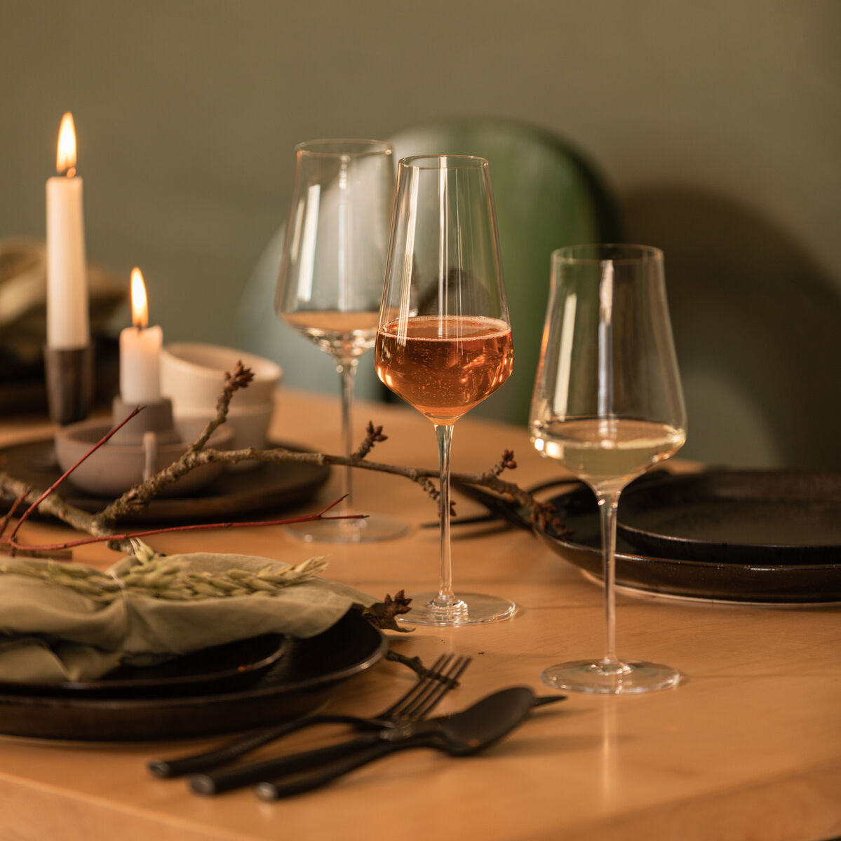Ein gedeckter Tisch mit zwei Weißweingläsern aus dem Set »DUO«. Neben den Gläsern befinden sich schwarze Teller, grüne Stoffservietten, Besteck und brennende Kerzen. Die Atmosphäre ist warm und gemütlich.
