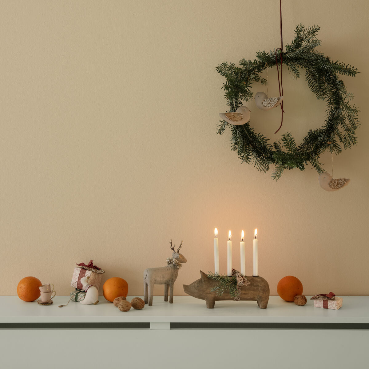 Großer Kerzenständer in Form eines Holzschweins mit vier brennenden Kerzen auf einer weißen Kommode, begleitet von weihnachtlichen Dekorationen wie einem Tannenzweig-Kranz, kleinen Holzfiguren, Nüssen und Mandarinen; stimmungsvolle, natürliche und festliche Atmosphäre.