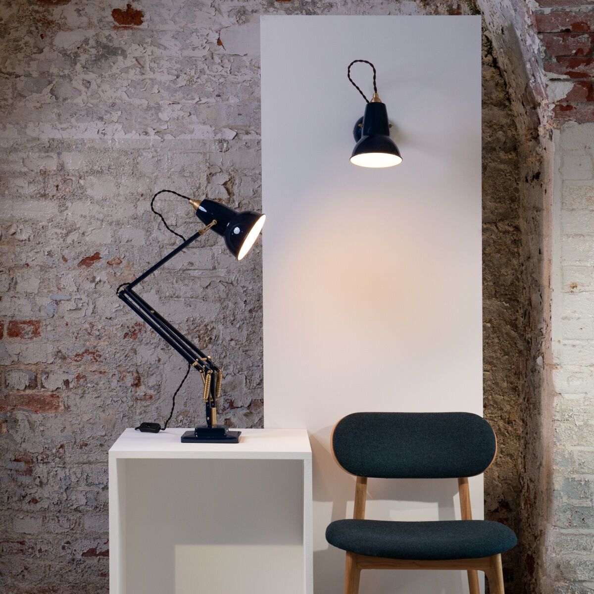 Eine Tischleuchte »Anglepoise Original 1227 Brass Desk Lamp« steht auf einem weißen Würfeltisch vor einer freiliegenden Ziegelwand. Eine weitere Lampe ist an einem weißen Wandpaneel montiert. Ein grünlich-grauer Stuhl mit Holzelementen ist rechts daneben platziert.
