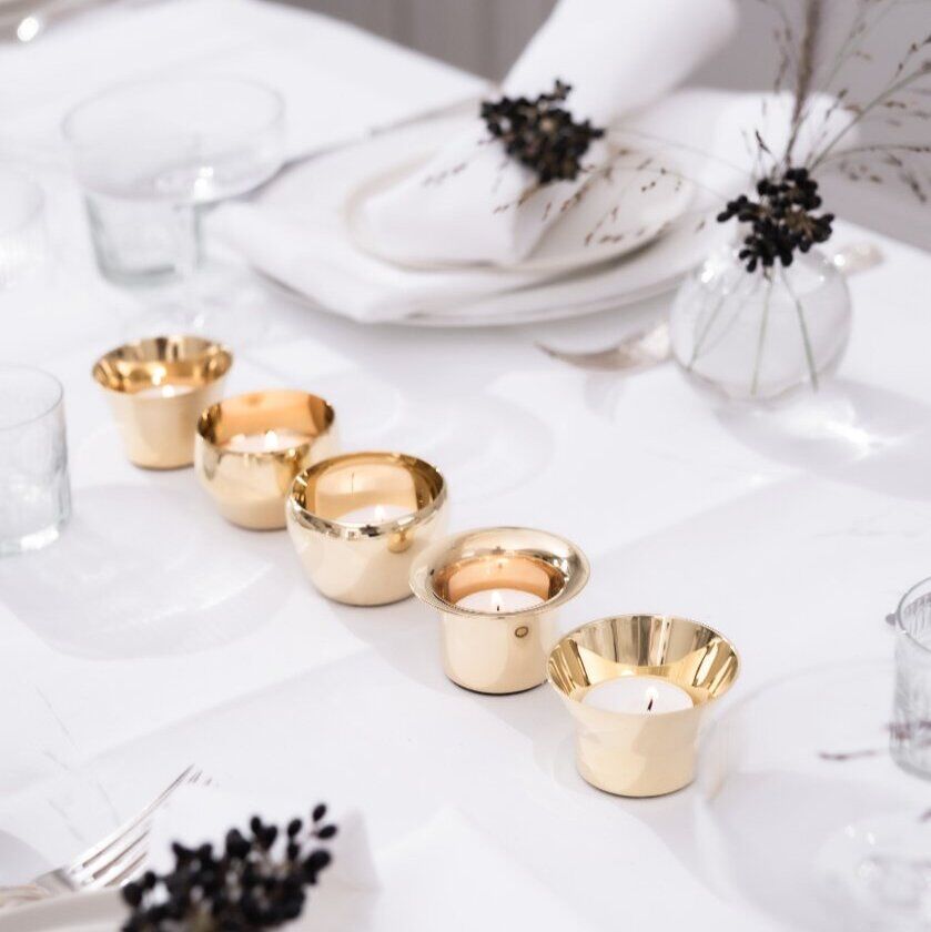 Ein elegantes Teelichter-Set aus goldenen Kerzenhaltern auf einem weiß gedeckten Tisch. Die Tischdekoration umfasst weiße Stoffservietten und kleine Blumenarrangements in Glasvasen, die für eine festliche Atmosphäre sorgen.