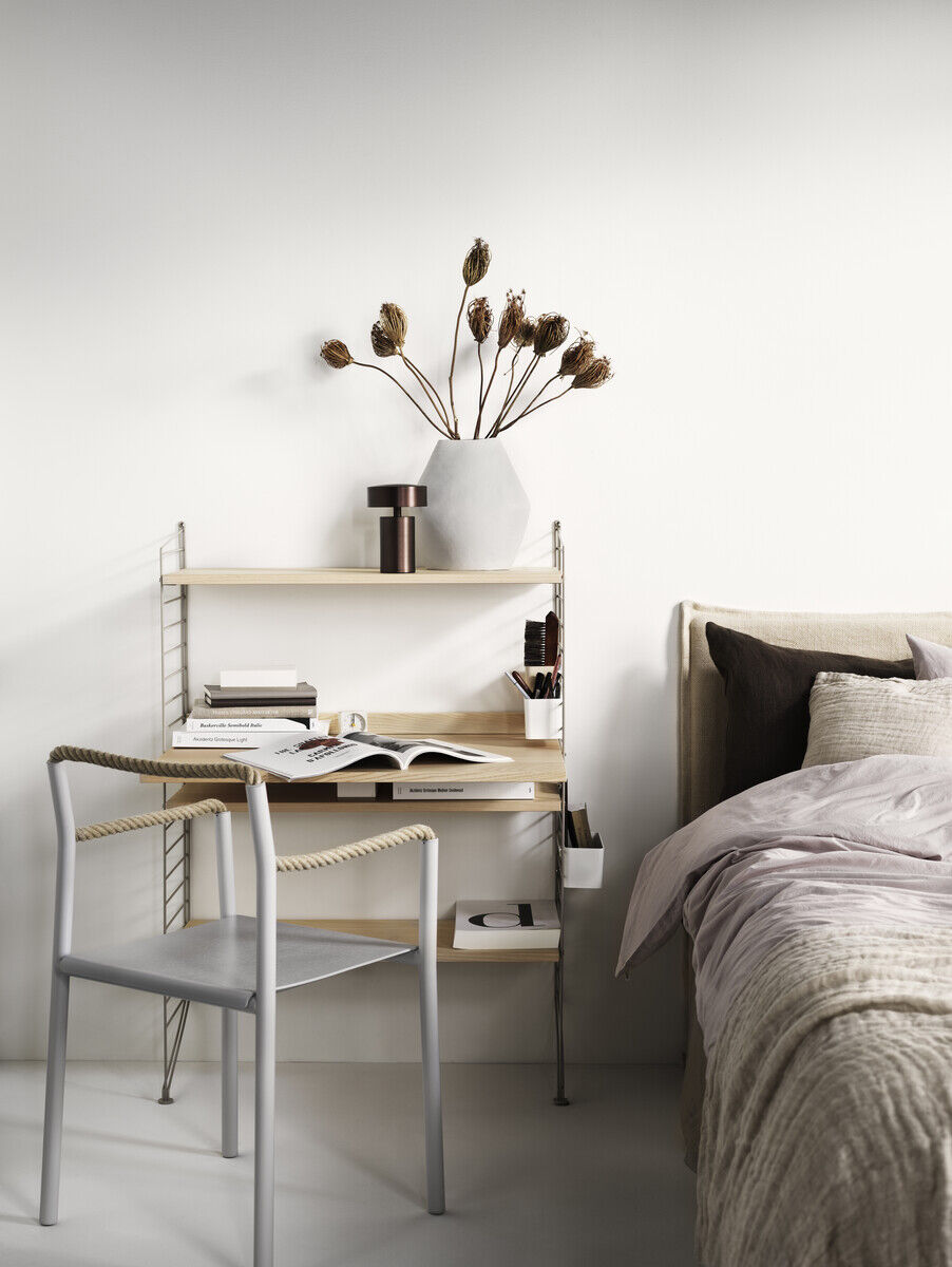 Ein minimalistischer Arbeitsplatz bestehend aus einem schlanken, modularen String-Möbelsystem mit Regalen, einer Arbeitsfläche und einer Metallstuhl mit einem Kissen. Der Schreibtisch ist in einem modernen, schlichten Design, das perfekt zu einem hellen, gut organisierten Raum passt. Neben dem Arbeitsplatz befindet sich ein schlichtes Bett. Auf dem Schreibtisch stehen Bücher, Schreibutensilien und eine Vase mit getrockneten Blumen. Die Farben im Bild sind Weiß, Beige, Grautöne und verschiedene Holztöne.