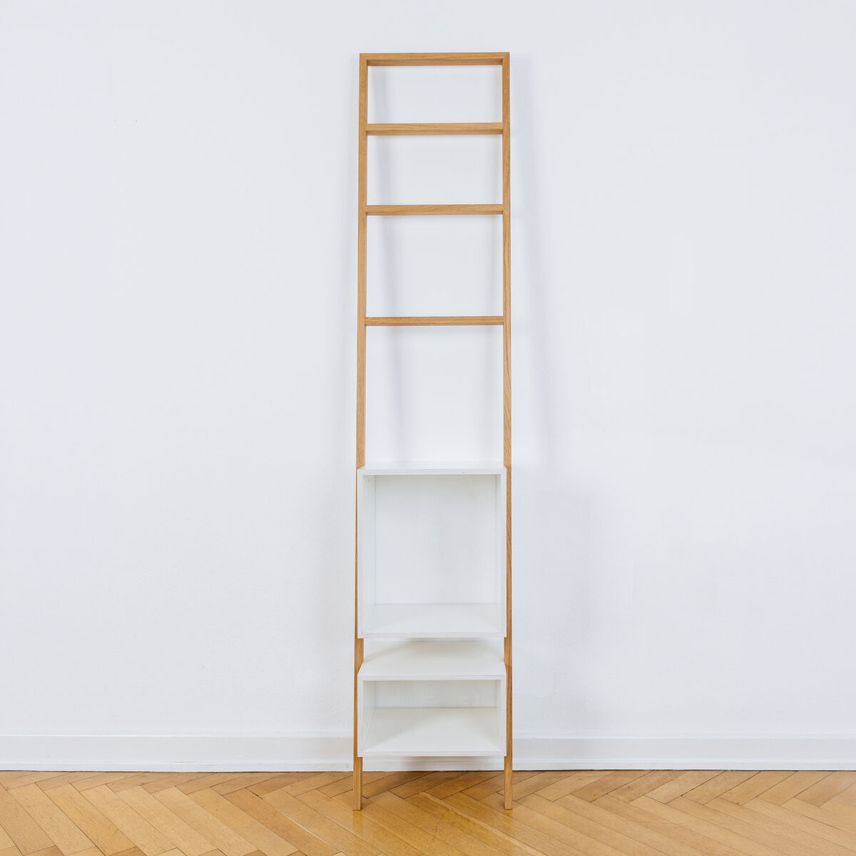Modernes Leiterregal »brecht« von das kleine b, aus geölter Eiche und weiß beschichteten Regalkästen. Das schlanke Regal lehnt an einer weißen Wand auf Parkettboden, bietet offene Fächer und kombiniert minimalistisches Design mit hoher Funktionalität.