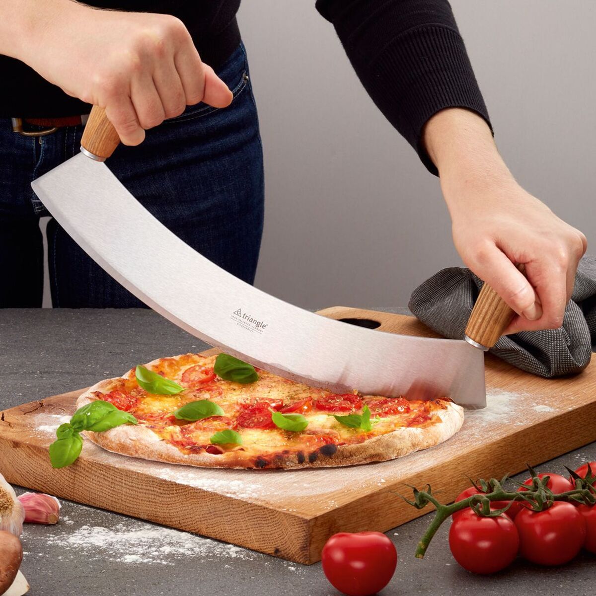 Eine Person schneidet mit einem einschneidigen Wiegemesser aus Esche Kernholz der Marke triangle eine Pizza auf einem Holzbrett. Auf der Pizza liegt frisches Basilikum. Im Vordergrund liegen Tomaten, im Hintergrund ein Küchentuch und Knoblauch auf einer anthrazitfarbenen Arbeitsfläche.