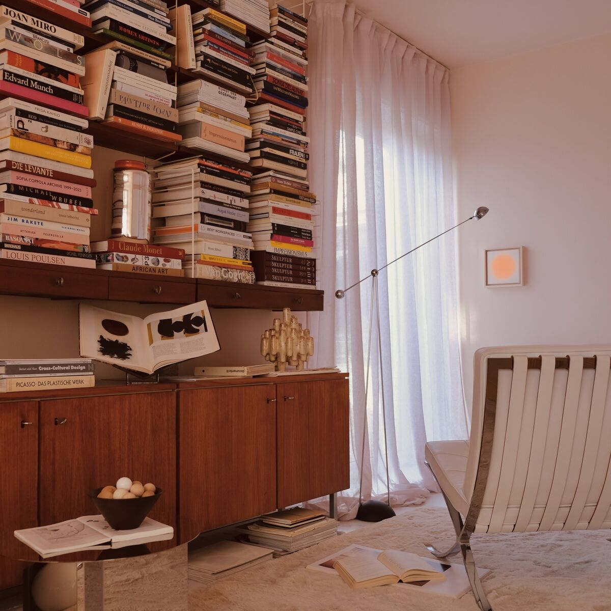 Modern eingerichtetes Wohnzimmer mit einem quadratischen, orangefarbenen Kunstwerk der ZEIT-Sonderedition Sorbet »Orange« (20x20cm) an der Wand, daneben ein Regal mit vielen Büchern, eine Designerliege aus weißem Leder, ein Holzmöbel und sanftes Tageslicht durch transparente Vorhänge.