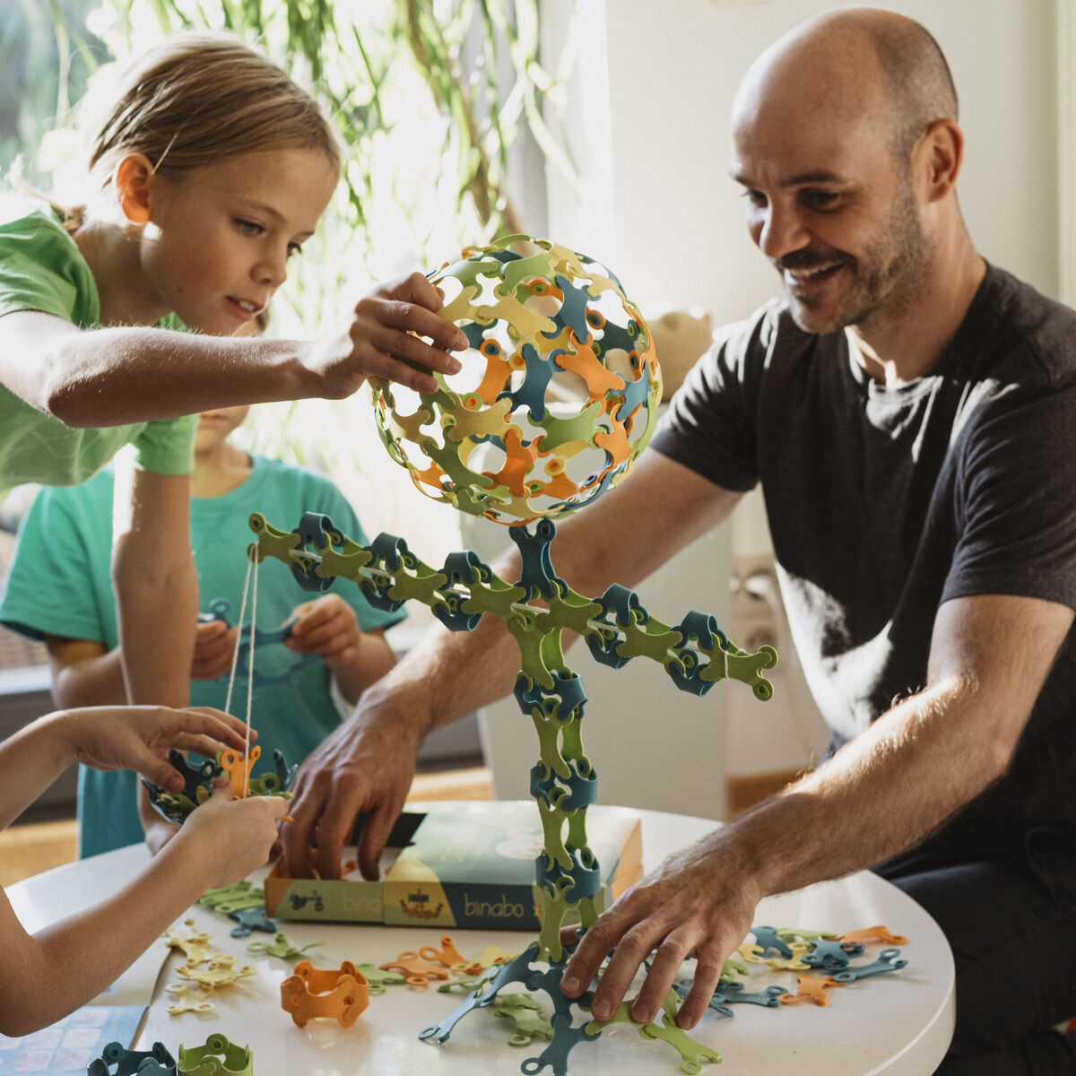Ein Mann und Kinder sitzen gemeinsam an einem Tisch und spielen mit Binabo, einem Kreativspielzeug aus bunten, zusammensteckbaren Teilen. Holzspielzeug steht auf dem Tisch.